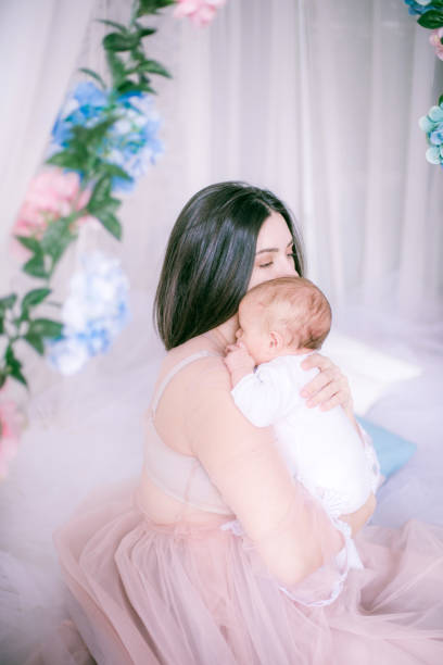 Young mother in a boudoir dress with a baby in her arms in spring flowers stock photo