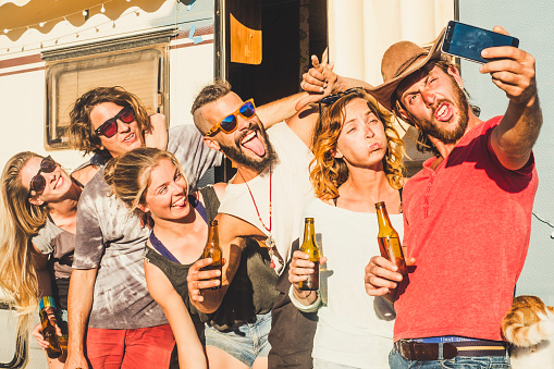 Group of crazy young caucasian people men and women taking selfie pictures doing funny expressions - friendship and youthful concept - old caravan in background and summer season