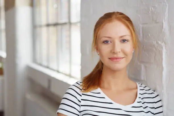 Thoughtful pretty young redhead woman in a striped t-shirt leaning on a white painted brick interior wall looking at the camera with a quiet smile