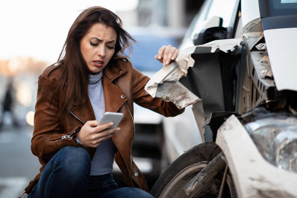 istock Young woman typing text message on mobile phone next to her wrecked car. 1135829248