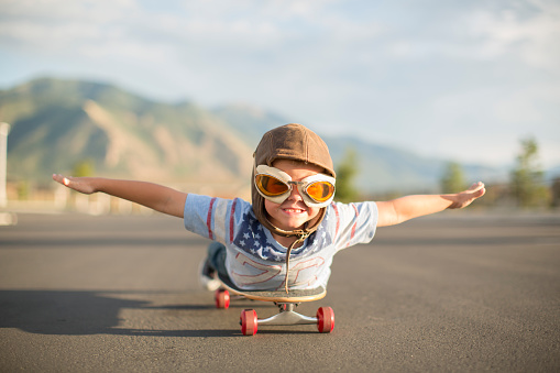 A young boy wearing a flight cap and goggles spreads his arms out dreaming of flying while lying down on a skateboard. He has an excited expression on his face as he aspires to be a pilot flying an airplane when he grows up. Image taken in Utah, USA.