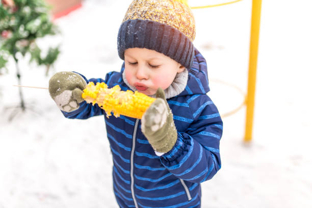 ein kleiner junge von 4-5 jahren isst im winter gekochten mais auf der straße, vor dem hintergrund von schnee in warmer jacke und hut, weht auf heißem mais, versucht, in der natur zu kühlen und zu kochen. - child winter snow 4 5 years stock-fotos und bilder