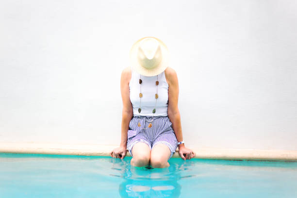 senior attivo: donna seduta al bordo della piscina, gambe in acqua - women wading sun hat summer foto e immagini stock