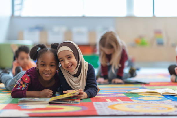 Reading Together Two girls are laying on the carpet in their classroom. They are smiling at the camera while reading a book. pics of a cute muslim outfits stock pictures, royalty-free photos & images