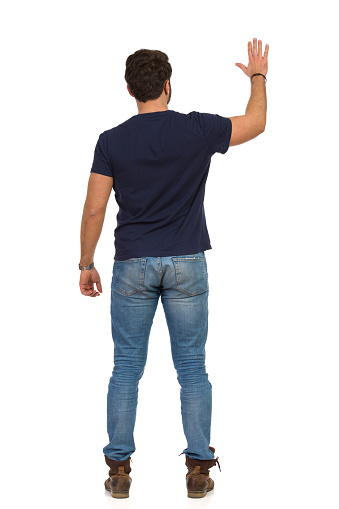 Man in jeans and blue t-shirt is standing with arm raised and waving hand. Rear view. Full length studio shot isolated on white.