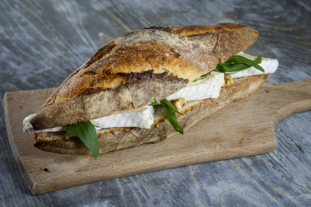 Brie sanwdich in a French baguette, made of Brie de Meaux Cheese with some slices of rucola salad on display on a rustic wooden table. This sandwich, in France, is called sandwich au brie stock photo