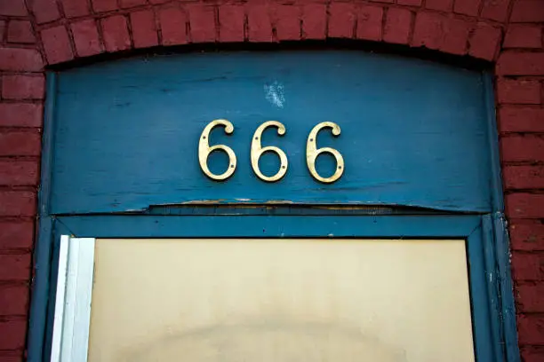The brass street address number '666' above a doorway on blue painted wood, framed by red brick.