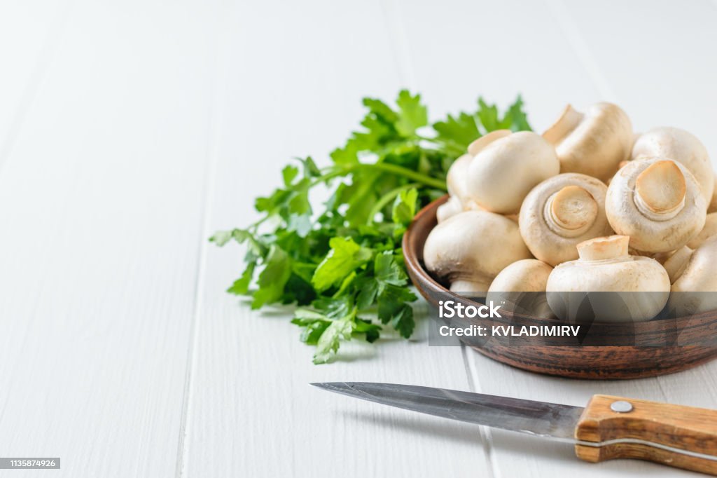 A knife next to a bowl of mushrooms and a bunch of parsley on a white table. Vegetarian cuisine. A knife next to a bowl of mushrooms and a bunch of parsley on a white table. Vegetarian cuisine. Natural plant food. Agaricus Stock Photo