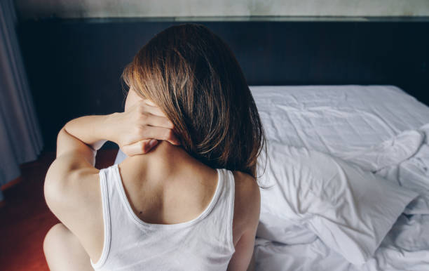 ベッドの上に座っている若いアジアの女性の背中のビューは、首の痛みを持ち、朝目覚めた後、彼女の体を伸ばします。 - neck pain ストックフォトと画像