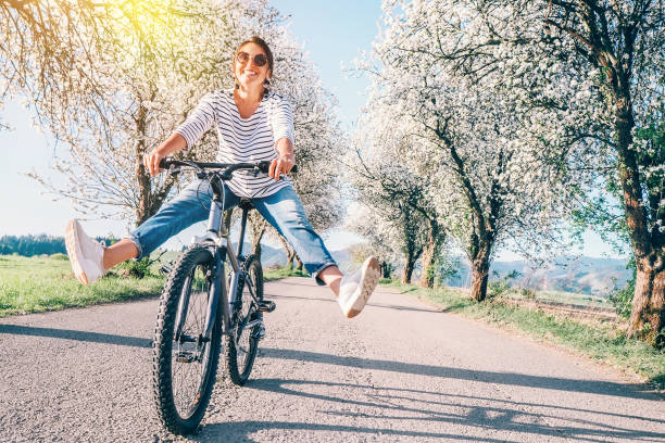 feliz sonriente mujer se extiende alegremente piernas en bicicleta en la carretera del país bajo árboles de flor. la primavera se acerca a la imagen conceptual. - comming fotografías e imágenes de stock