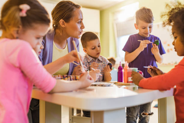 istock Young preschool teacher coloring Easter eggs with group of kids. 1135906029