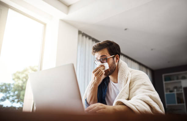The flu has got him working from home today Shot of a sickly young businessman blowing his nose with a tissue while working from home cold and flu man stock pictures, royalty-free photos & images