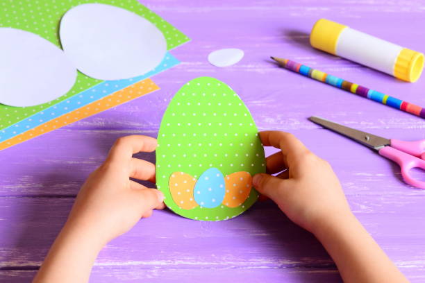 istock Small child holding a Easter card in hands. Child made Easter greeting card in egg shape. Materials for art creativity on a wooden table. Paper crafts activity in kindergarten or at home 1135912645