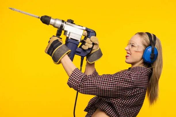 Beautiful young woman in goggles and headphones worth holding perforator drill isolated on yellow background - Image