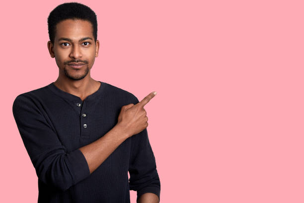 istock Handsome young african man dresses black shirt, points aside with his right hand. Good looking male shows somethig interesting to his friends. Model poses in studio against pink wall. People concept. 1135915472