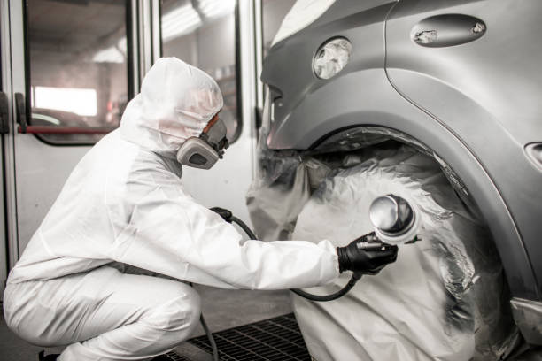 Car body shop technician in full coveralls and a breathing mask applying a coat of paint on a car part in a painting booth Painting technician applying a coat of paint on a car segment in a body shop painting chamber. door panel stock pictures, royalty-free photos & images