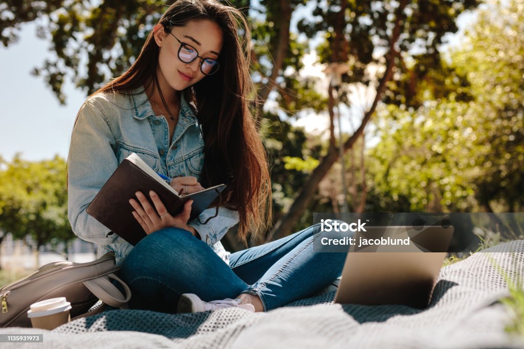 Young woman studying sitting outdoors Close up of a college student sitting in a park and writing in her notebook. College student making notes looking at her laptop computer. Student Stock Photo Young woman studying sitting outdoors Close up of a college student sitting in a park and writing in her notebook. College student making notes looking at her laptop computer. Student Stock Photo
