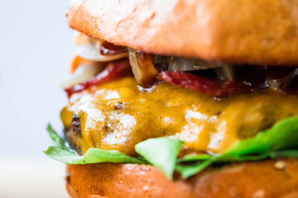 Close up image of a freshly flame grilled beef burger loaded with melted cheese and crispy bacon with fresh green salad leaves, sandwiched between a toasted sesame seed bun. White backgroung with room for copy space.
