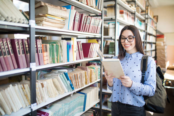 istock Young student with books and backpack standing by bookshelf in college library 1135938749