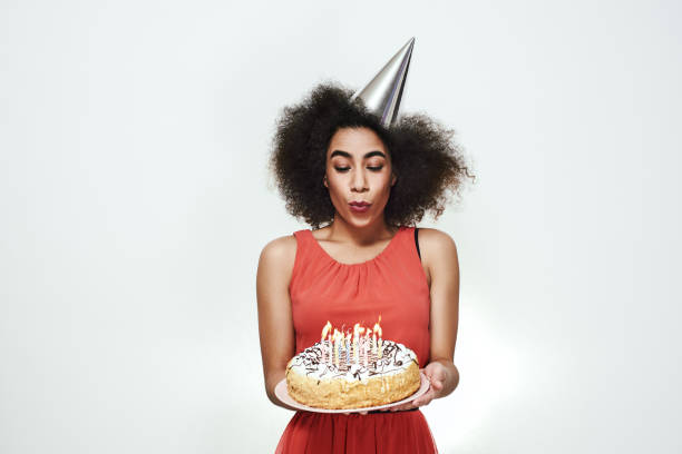 シルバーパーティーハットでかなり若いアフロアメリカの女性は彼女の誕生日を祝っているとおいしいケーキのろうそくを吹き飛ばす - cake birthday candle blowing ストックフォトと画像