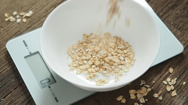 Product weighing concept. Man pours oatmeal into a white bowl on the kitchen scales on wooden background