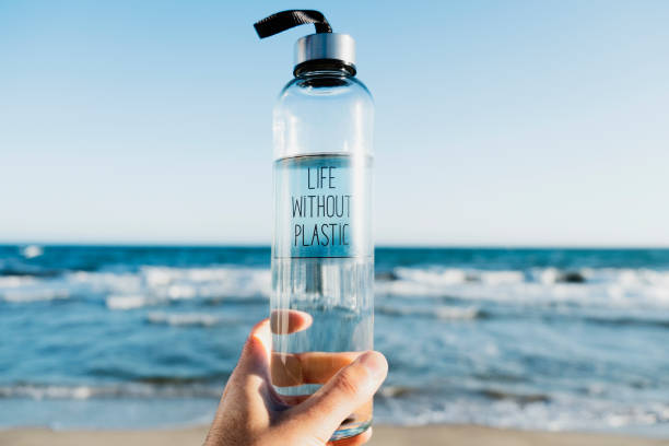 water bottle with the text life without plastic closeup of a caucasian man holding a glass reusable water bottle with the text life without plastic written in it, on the beach, with the ocean in the background purified water photos stock pictures, royalty-free photos & images