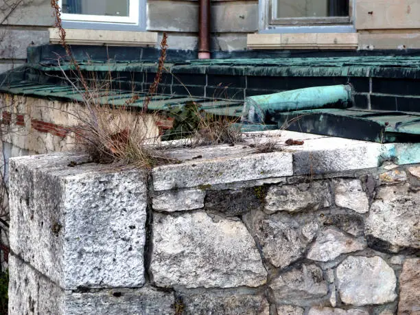 carved and shaped textured stone wall and ledge detail with green aged copper flashing, roof and rain water leader. white window sill and dry weed on the ledge.