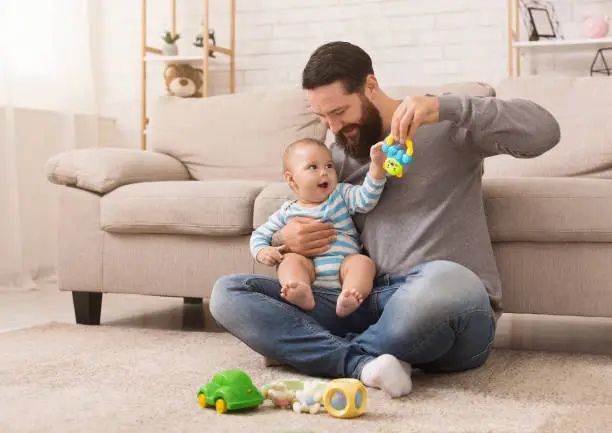 Happy father playing with his baby son with bright rattle, sitting on floor at home, copy space