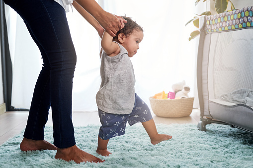 Baby's first steps holding mother's hands, cute unstable walking in home nursery with cot