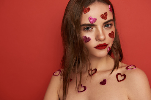 Cropped photo of charming young lady looking at camera in studio. Her face is covered with decorative glossy hearts. Close up. Love concept