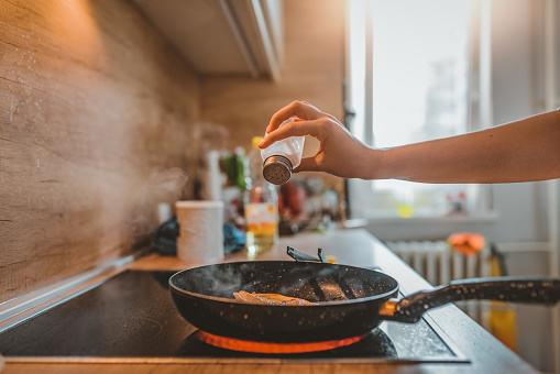 Photo of Unrecognizable woman Adding salt. Back light to salt from salt shaker. Close up. Young Caucasian lady standing in the kitchen while adding salt to the meal in frying pan with chicken breast.
