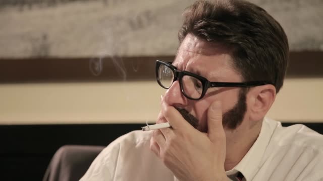Men with mustaches and glasses smokes cigarette while sitting on a desk