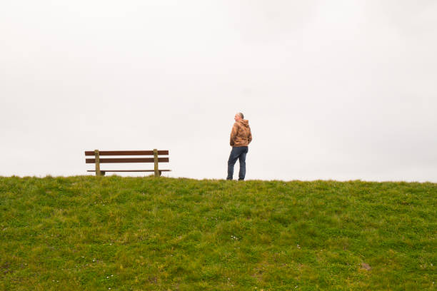 A single person standing next to a single empty wooden bench on the horizon above a grass field against a large grey clouded sky depicting loneliness, thinking, pondering, grieve, being alone, sadness