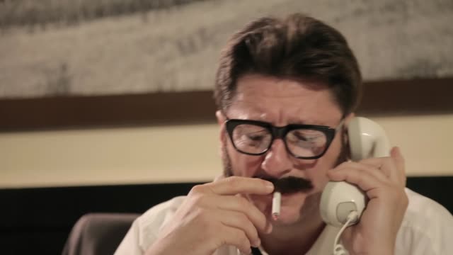 Men with mustaches and glasses smokes cigarette, talking on a retro phone and smiling, while sitting on a desk