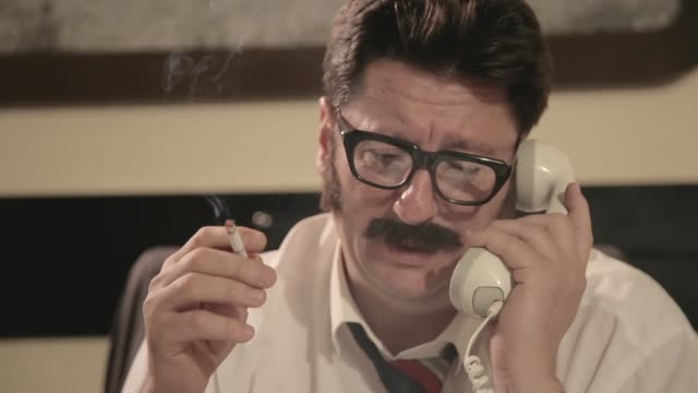 Men with mustaches and glasses smokes cigarette, talking on a retro phone, while sitting on a desk