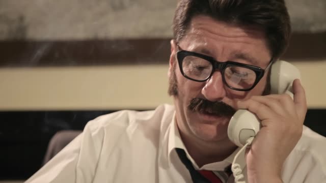 Men with mustaches and glasses smokes cigarette, talking on a retro phone, while sitting on a desk