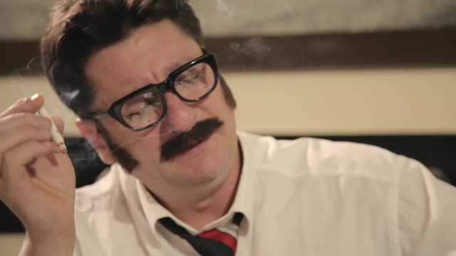 Men with mustaches and glasses smokes cigarette, hanging up a phone, while sitting on a desk