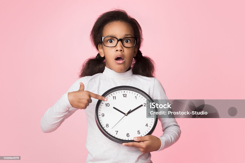 Surprised african-american girl holding big clock and pointing on it Surprised african-american girl holding big clock and pointing on it over pink background Child Stock Photo
