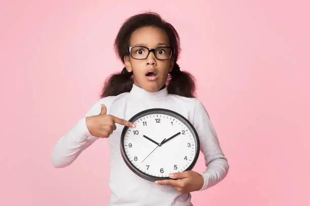 Photo of Surprised african-american girl holding big clock and pointing on it