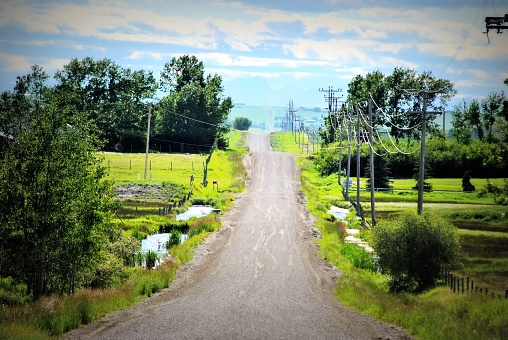 An unusual looking rural road in the country.  The road leads upward toward the clouds and beyond. The shallow ditches are  filled with water.  Trees and green grass are on both sides of the road.