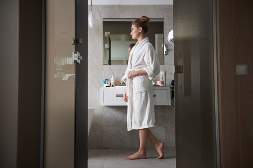 Self care and hygiene concept. Side on full length portrait of young smiling woman in white bathrobe staying in bathroom near wash basin
