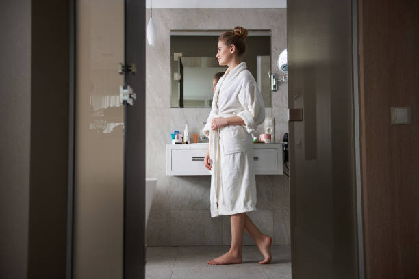 Young careful smiling lady staying in bathroom Self care and hygiene concept. Side on full length portrait of young smiling woman in white bathrobe staying in bathroom near wash basin bathrobe stock pictures, royalty-free photos & images