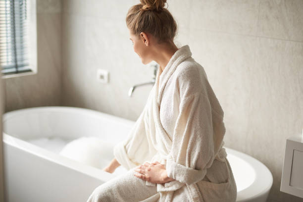 Young lady in bathrobe ready taking bath Self care treatment concept. Waist up side on portrait of young woman sitting on tub and touching foam water before taking bath bathtub stock pictures, royalty-free photos & images