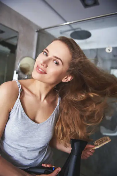 Happy in ordinary thing. Close up portrait of cheerful woman enjoying while drying her voluminous hair by hairdryer in bathroom