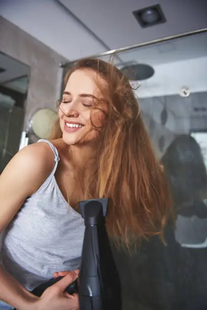 Happy in ordinary thing. Close up portrait of cheerful charming woman enjoying of drying her voluminous hair in bathroom