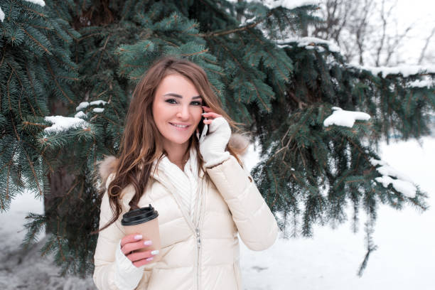 ragazza bruna si trova in inverno sullo sfondo del parco di alberi di natale innevati. telefonate felici sorrisi parlare. nella sua mano tiene una tazza di tè caldo al caffè. applicazione online su internet, ascolto audio. - tourist resort audio foto e immagini stock