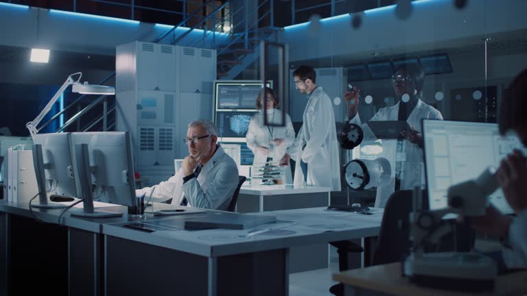 Diverse International Team of Industrial Scientists and Engineers Wearing White Coats Working on Heavy Machinery Design in Research Laboratory. Professionals Using Computers and Talking