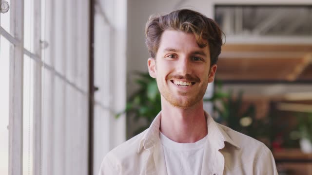 Millennial white male creative standing in office, turning and smiling to camera, close up