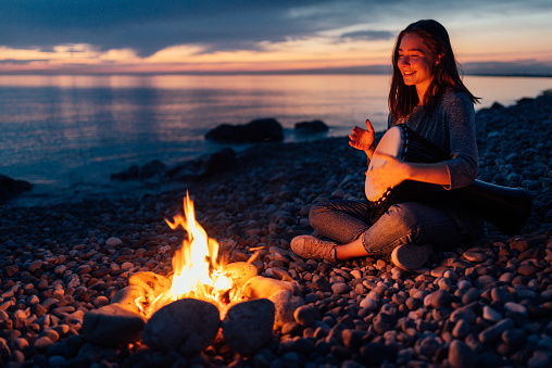 cheerful percussionist girl playing djembe sitting on the beach by the fire at sunset.