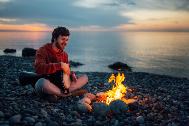 istock percussionist guy plays a djembe sitting on beach by the fire at sunset. 1135995203